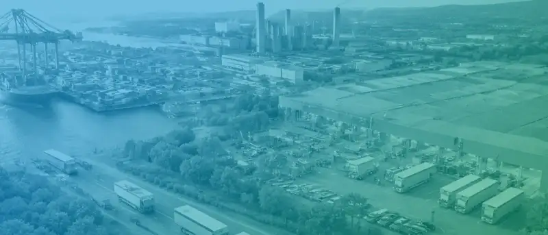 Luftaufnahme eines Industriehafens mit Containern, Kränen, Lagerhallen und mehreren Lkw auf einer Straße, blau-grün eingefärbt.