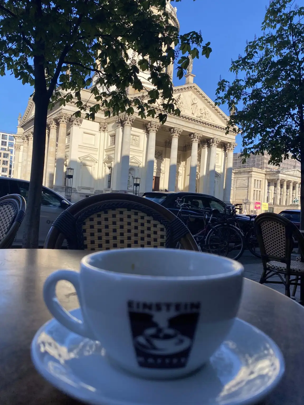 Eine Kaffeetasse auf einem Außentisch vor einem historischen Gebäude in der Sonne.