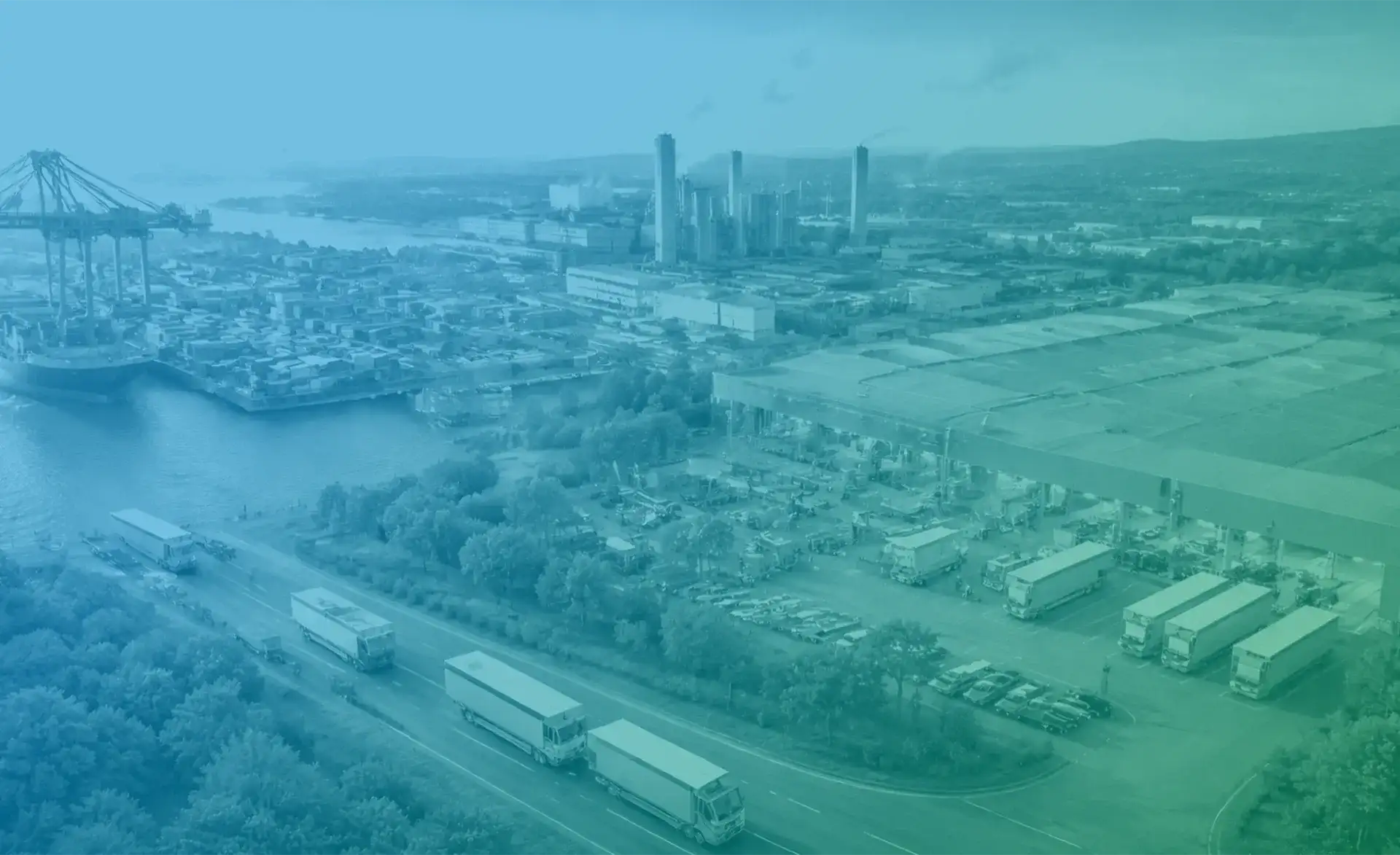 Luftaufnahme eines Industriehafens mit Containern, Kränen, Lagerhallen und mehreren Lkw auf einer Straße, blau-grün eingefärbt.