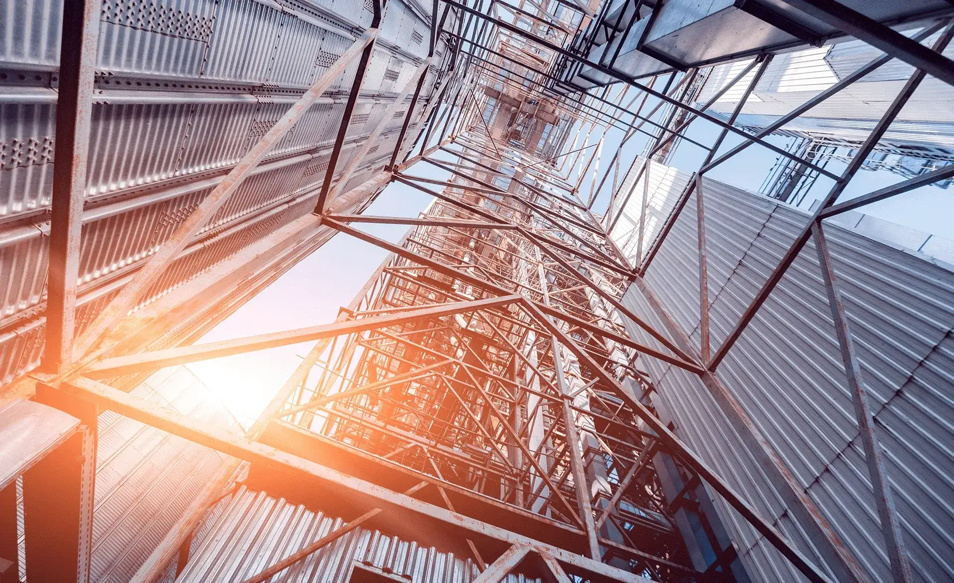 Blick nach oben durch ein Gerüst aus Stahlträgern zwischen hohen Industriegebäuden bei Sonnenlicht.