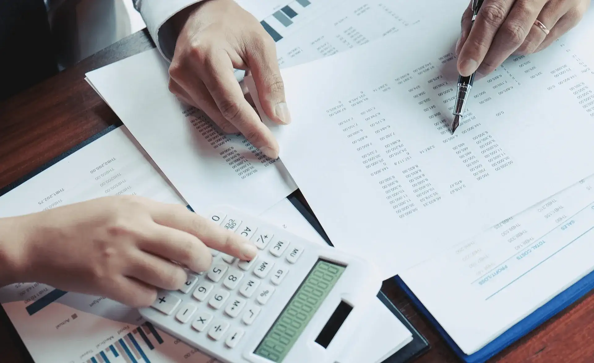 Zwei Personen analysieren Finanzunterlagen; eine Hand nutzt einen Taschenrechner, die andere zeigt auf Tabellen.