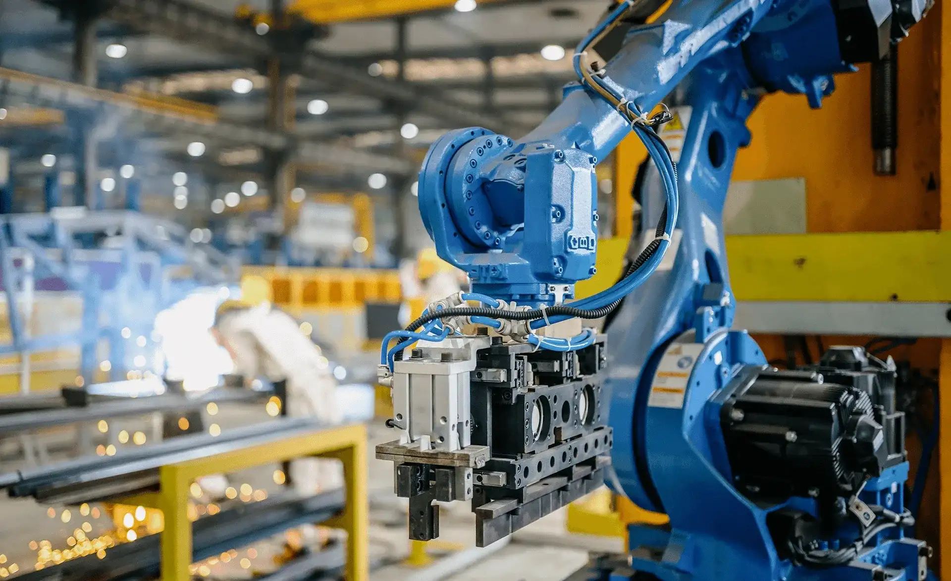 Industrieroboterarm arbeitet in einer Produktionshalle, während im Hintergrund ein Arbeiter Funken schweißt.